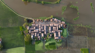 Aerial view of a rural village with tightly clustered, rectangular buildings surrounded by lush green fields and a large water body. The fields show a patchwork of different shades of green, and the water appears calm with some vegetation.