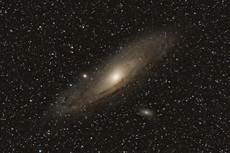 An expansive view of a galaxy surrounded by numerous stars. The galaxy has a bright, centralized core and extends outward with a diffuse, hazy appearance, showcasing its spiral structure. The background is filled with a rich scattering of stars against the blackness of space.