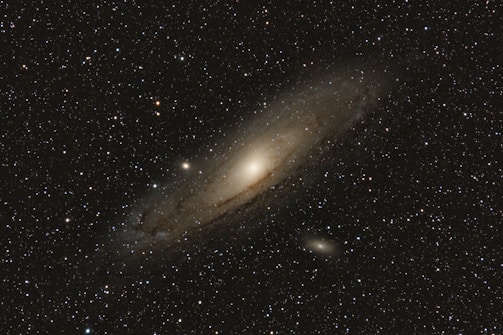An expansive view of a galaxy surrounded by numerous stars. The galaxy has a bright, centralized core and extends outward with a diffuse, hazy appearance, showcasing its spiral structure. The background is filled with a rich scattering of stars against the blackness of space.