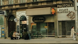 A city street scene featuring a facade of shops with vintage signage, including Nikon and Kodak, suggesting a camera store. A pair of people sit outside at a small cafe on wooden benches, engaged in conversation. A bicycle is parked against the building, and a person in a long coat walks by.