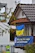 A building facade features a banner with German text and an image of a person's face. Below, there is a blue and yellow Ukrainian flag hanging from the building. The architecture includes a roof with weathered tiles and a small window. In the foreground, branches with leaves are partially in view, and a sign is visible near a fence.