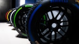 A row of Pirelli Formula 1 tires is lined up, each displaying a colorful marking indicating different compounds. The tires are set on a smooth white surface with a dark, industrial background.