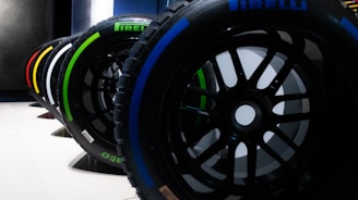 A row of Pirelli Formula 1 tires is lined up, each displaying a colorful marking indicating different compounds. The tires are set on a smooth white surface with a dark, industrial background.