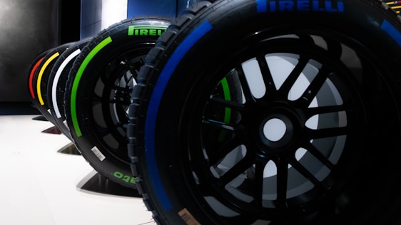 A row of Pirelli Formula 1 tires is lined up, each displaying a colorful marking indicating different compounds. The tires are set on a smooth white surface with a dark, industrial background.