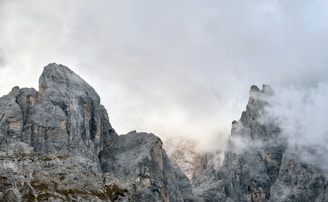 A rugged mountain range with jagged peaks partially shrouded in dense mist. The rocky surface features shades of gray and brown, with some vegetation visible on the lower slopes. The atmosphere is serene and mystical, with a layer of clouds obscuring parts of the mountains.