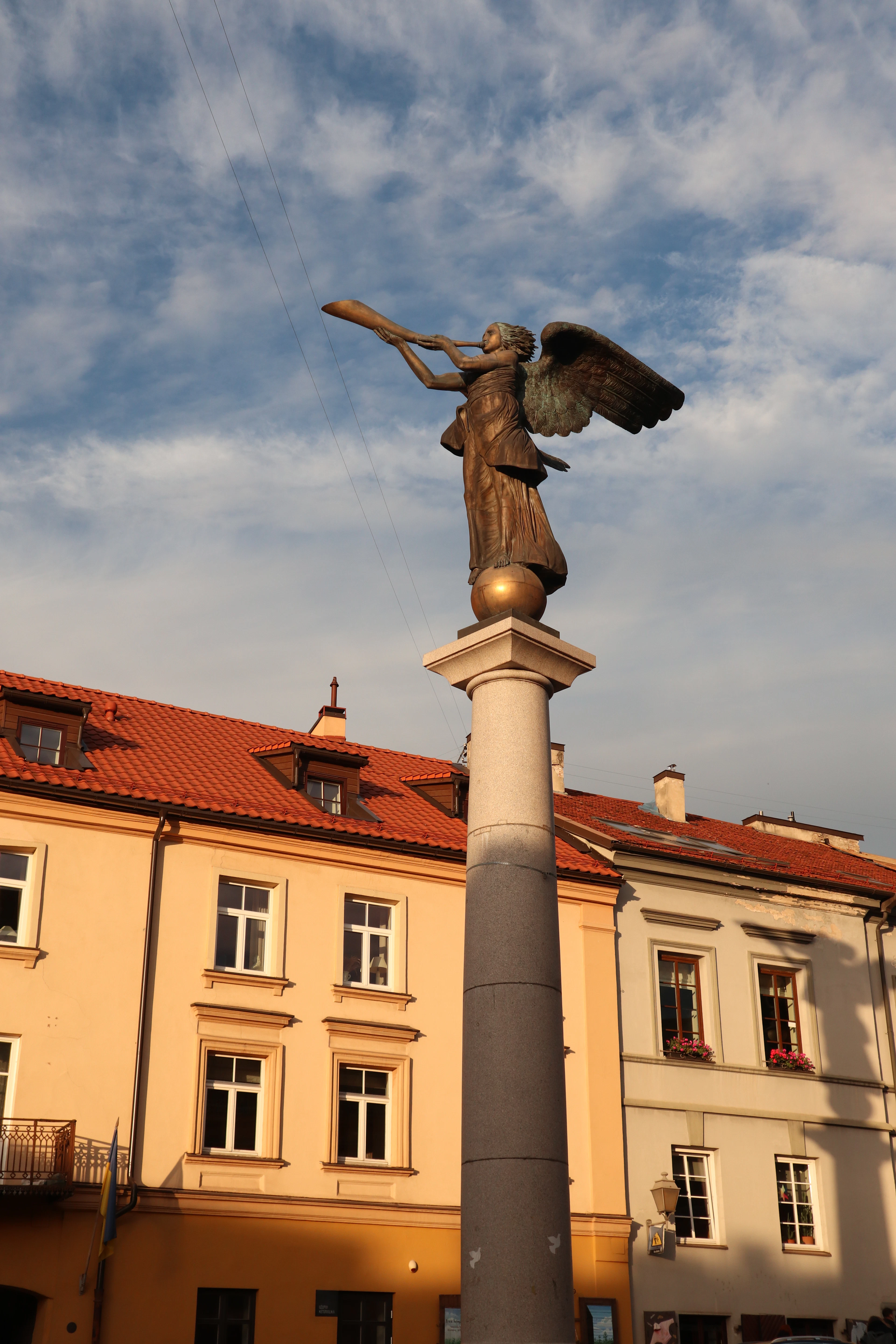The bronze Angel of Užupis blowing his trumpet from the top of his stone column in Vilnius, gilded by low evening sun above the ochre walls and red-tiled roofs of the district.
