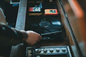 A person adjusts a vinyl record on a vintage turntable. The setup includes vinyl records, some 8-track tapes, and knobs for adjusting sound levels. The atmosphere suggests a retro or nostalgic vibe.