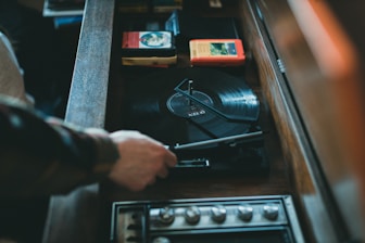 A person adjusts a vinyl record on a vintage turntable. The setup includes vinyl records, some 8-track tapes, and knobs for adjusting sound levels. The atmosphere suggests a retro or nostalgic vibe.
