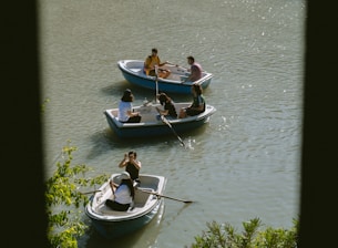 Several small rowboats are floating on a calm body of water. The boats each contain a group of people who seem relaxed and engaged in conversation. The sunlight glistens on the water, and green foliage is visible in the foreground.
