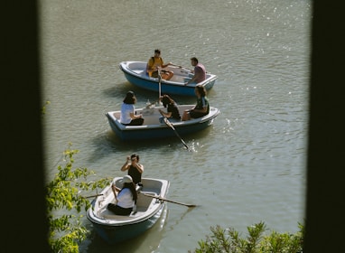Several small rowboats are floating on a calm body of water. The boats each contain a group of people who seem relaxed and engaged in conversation. The sunlight glistens on the water, and green foliage is visible in the foreground.