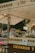 A bustling churro stand with a variety of snacks displayed, including churros and bottled drinks. The vendor is preparing or selling items at the counter under a canopy. There are decorative items, including flowers or plants, and people are visible in the background, suggesting a lively outdoor setting.