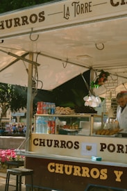 A bustling churro stand with a variety of snacks displayed, including churros and bottled drinks. The vendor is preparing or selling items at the counter under a canopy. There are decorative items, including flowers or plants, and people are visible in the background, suggesting a lively outdoor setting.