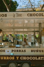 A street food cart with the name 'La Torre' is selling churros and other snacks. The cart has signage displaying the words 'churros', 'porras', and 'chocolate'. A person wearing a white shirt is inside the cart, surrounded by cooking equipment and food displays. There are some trees visible in the background, suggesting an outdoor setting.