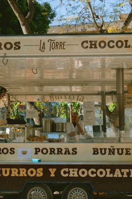 A street food cart with the name 'La Torre' is selling churros and other snacks. The cart has signage displaying the words 'churros', 'porras', and 'chocolate'. A person wearing a white shirt is inside the cart, surrounded by cooking equipment and food displays. There are some trees visible in the background, suggesting an outdoor setting.