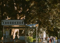 A churros stand is situated under a large, leafy tree. The stand has a sign reading 'Churreria' and is adorned with various items including bottles of water and kitchen equipment. Surrounding it is a bustling park scene with numerous people walking along a shaded path, enjoying a sunny day.