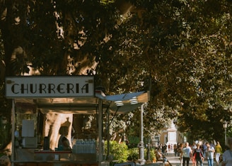 A churros stand is situated under a large, leafy tree. The stand has a sign reading 'Churreria' and is adorned with various items including bottles of water and kitchen equipment. Surrounding it is a bustling park scene with numerous people walking along a shaded path, enjoying a sunny day.