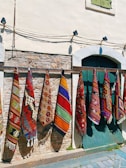 Colorful woven rugs with intricate geometric patterns hang vertically on a wooden rail against a rustic stone and plaster wall. A window with green shutters is partially visible above, and cobbled stones lie at the base.