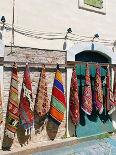 Colorful woven rugs with intricate geometric patterns hang vertically on a wooden rail against a rustic stone and plaster wall. A window with green shutters is partially visible above, and cobbled stones lie at the base.