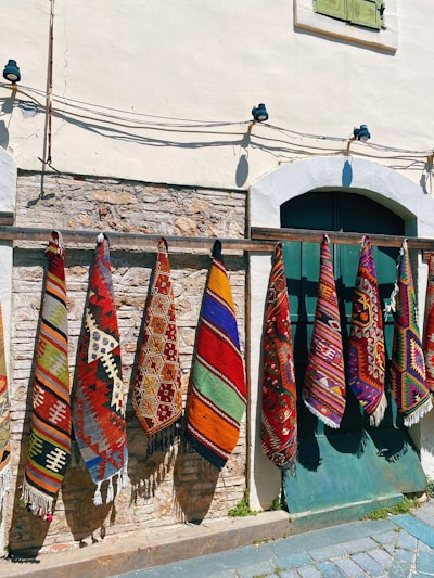 Colorful woven rugs with intricate geometric patterns hang vertically on a wooden rail against a rustic stone and plaster wall. A window with green shutters is partially visible above, and cobbled stones lie at the base.