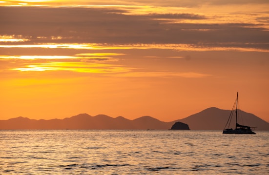 A serene seascape with calm waters under a vibrant orange and yellow sunset. The silhouette of distant islands is visible on the horizon, with a sailboat gracefully floating in the foreground. The sky is filled with streaks of orange, yellow, and subtle hints of red, casting a reflective glow on the water.