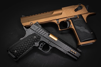 Two handguns are arranged on a dark surface. One is a dark colored pistol with a honeycomb-patterned grip and gold barrel, while the other is a bronze-colored pistol with a textured grip.