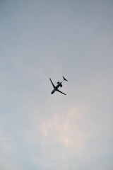 A passenger airplane is captured flying overhead against a backdrop of a cloudy sky. The aircraft is prominently centered, with its wings and tail clearly visible. The sky is a light blue-gray, subtly transitioning to patches of soft white clouds.
