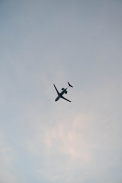 A passenger airplane is captured flying overhead against a backdrop of a cloudy sky. The aircraft is prominently centered, with its wings and tail clearly visible. The sky is a light blue-gray, subtly transitioning to patches of soft white clouds.