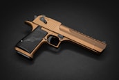 A metallic, bronze-colored handgun is positioned at an angle on a dark background. The grip is textured and black, contrasting with the bronze finish of the weapon. The overall design features sleek lines and curves, with visible details such as the safety switch and trigger.
