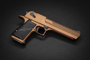 A metallic, bronze-colored handgun is positioned at an angle on a dark background. The grip is textured and black, contrasting with the bronze finish of the weapon. The overall design features sleek lines and curves, with visible details such as the safety switch and trigger.