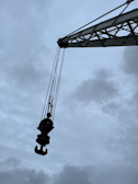 A large crane arm extends diagonally across the image with a heavy-duty hook hanging from it, silhouetted against a cloudy, overcast sky. The industrial equipment appears robust and weathered, highlighting its use in construction or lifting tasks.