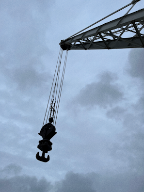 A large crane arm extends diagonally across the image with a heavy-duty hook hanging from it, silhouetted against a cloudy, overcast sky. The industrial equipment appears robust and weathered, highlighting its use in construction or lifting tasks.