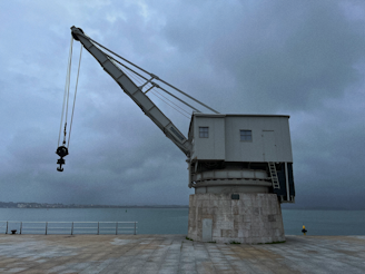 A large industrial crane is positioned on a concrete platform by the edge of a body of water. The sky is overcast, giving the scene a moody, subdued atmosphere. The water is calm and there is a faint silhouette of a shoreline in the distance.