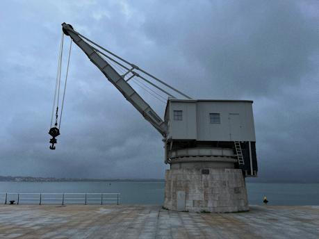A large industrial crane is positioned on a concrete platform by the edge of a body of water. The sky is overcast, giving the scene a moody, subdued atmosphere. The water is calm and there is a faint silhouette of a shoreline in the distance.