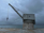 A large industrial crane is positioned on a concrete platform by the edge of a body of water. The sky is overcast, giving the scene a moody, subdued atmosphere. The water is calm and there is a faint silhouette of a shoreline in the distance.