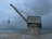 A large industrial crane is positioned on a concrete platform by the edge of a body of water. The sky is overcast, giving the scene a moody, subdued atmosphere. The water is calm and there is a faint silhouette of a shoreline in the distance.