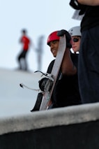 A person is holding a snowboard while wearing a red helmet. Another person stands nearby with a white helmet and sunglasses. The background shows a blurred figure in motion wearing a red shirt.