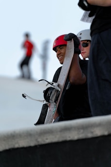 A person is holding a snowboard while wearing a red helmet. Another person stands nearby with a white helmet and sunglasses. The background shows a blurred figure in motion wearing a red shirt.