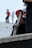 A person is holding a snowboard while wearing a red helmet. Another person stands nearby with a white helmet and sunglasses. The background shows a blurred figure in motion wearing a red shirt.