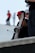 A person is holding a snowboard while wearing a red helmet. Another person stands nearby with a white helmet and sunglasses. The background shows a blurred figure in motion wearing a red shirt.