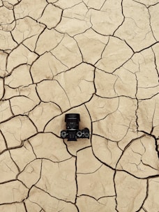A camera placed on cracked, dry earth. The ground is fractured into large, uneven pieces, showing signs of drought or arid conditions.