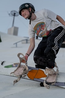 A person wearing a helmet and a white graphic t-shirt is engaged in a snowboarding activity. The individual appears to be in mid-action, possibly in a dynamic or freestyle move, with one hand gripping the snowboard. The expression is one of joy or excitement, reflecting the thrill of the sport. The background features a clear sky with some structures, suggesting this might be a snow park or a similar setup.