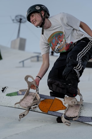 A person wearing a helmet and a white graphic t-shirt is engaged in a snowboarding activity. The individual appears to be in mid-action, possibly in a dynamic or freestyle move, with one hand gripping the snowboard. The expression is one of joy or excitement, reflecting the thrill of the sport. The background features a clear sky with some structures, suggesting this might be a snow park or a similar setup.