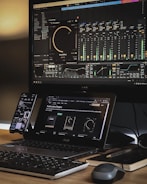 A workspace setup featuring a large monitor displaying audio mixing software with equalizer and sound effect controls. A laptop is placed beneath the monitor, showing a webpage about audio plugins. A smartphone is mounted to the side, and a keyboard and mouse rest on the wooden desk. The area is organized with a notepad and a pen nearby, creating an efficient and tech-focused environment.