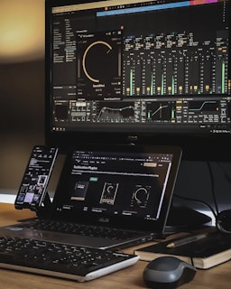 A workspace setup featuring a large monitor displaying audio mixing software with equalizer and sound effect controls. A laptop is placed beneath the monitor, showing a webpage about audio plugins. A smartphone is mounted to the side, and a keyboard and mouse rest on the wooden desk. The area is organized with a notepad and a pen nearby, creating an efficient and tech-focused environment.