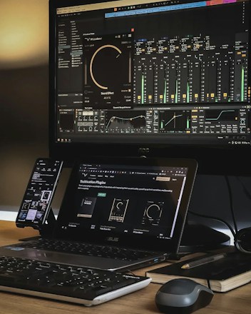 A workspace setup featuring a large monitor displaying audio mixing software with equalizer and sound effect controls. A laptop is placed beneath the monitor, showing a webpage about audio plugins. A smartphone is mounted to the side, and a keyboard and mouse rest on the wooden desk. The area is organized with a notepad and a pen nearby, creating an efficient and tech-focused environment.