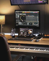 A music production workspace featuring a keyboard, laptop, smartphone, and two monitors displaying digital audio workstation software. The setting includes a wooden desk with a desk lamp, an audio interface, and a computer mouse.
