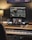 A music production workspace featuring a keyboard, laptop, smartphone, and two monitors displaying digital audio workstation software. The setting includes a wooden desk with a desk lamp, an audio interface, and a computer mouse.