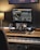A music production workspace featuring a keyboard, laptop, smartphone, and two monitors displaying digital audio workstation software. The setting includes a wooden desk with a desk lamp, an audio interface, and a computer mouse.