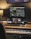 A music production workspace featuring a keyboard, laptop, smartphone, and two monitors displaying digital audio workstation software. The setting includes a wooden desk with a desk lamp, an audio interface, and a computer mouse.