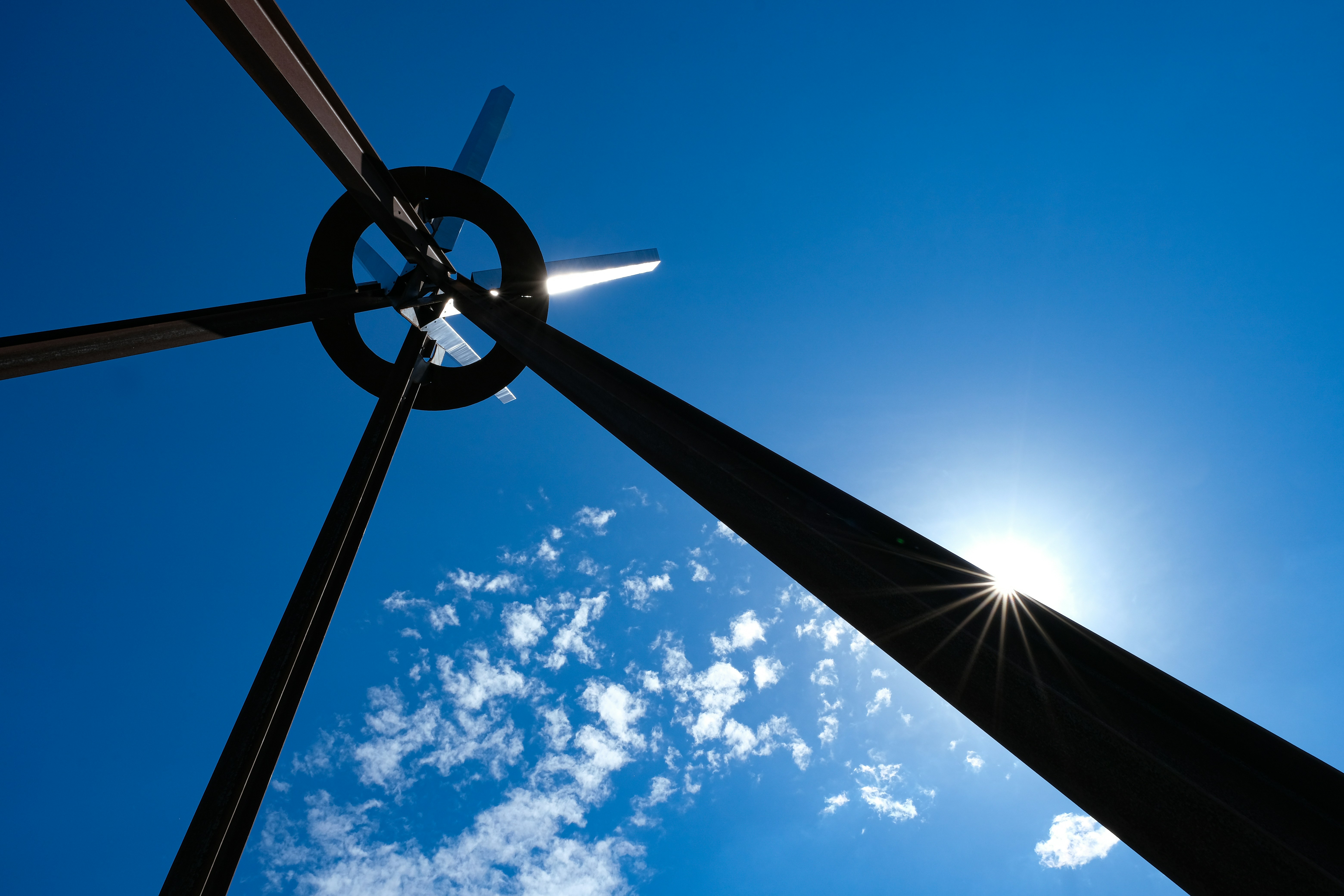Striking sculpture photographed from a low angle, set against a clear blue sky. It features bold, geometric shapes—a circular element at the center with radiating beams that suggest movement or rotation. The sun, positioned just above the sculpture, creates a brilliant flare effect, enhancing the dynamic and modern aesthetic of the artwork. The deep black of the sculpture contrasts vividly with the bright sky and soft, white clouds, drawing the viewer's attention to the interplay of light and shadow, and the precision of the sculpture's design.